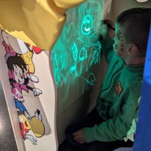 Kniender Junge blickt auf grün projizierte Meeresmotive an Wand; daneben ein bunter Comicstreifen und ein gelber Vorhang.