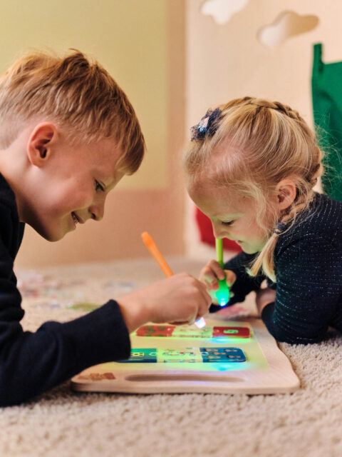 Zwei Kinder liegen auf dem Boden und legen bunte Holzbausteine auf ein Brett, konzentriert beim Spielen.