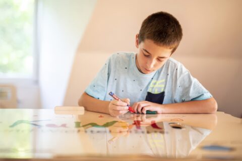 Junge zeichnet am Tisch mit bunten Stiften; Farbkasten und kindgerechtes Spielzeug auf dem Tisch.